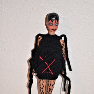 Peut inclure: Une poupée avec des cheveux noirs et du maquillage, vêtue d'un haut noir tricoté avec un design "X" rouge. Les jambes de la poupée sont peintes avec des lignes ondulées noires. De longues mèches noires s'étendent des épaules de la poupée.