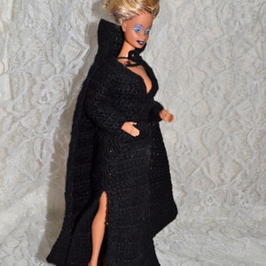 Puede incluir: Una muñeca con un vestido largo negro con capucha de ganchillo y una abertura alta. La muñeca tiene el pelo rubio y está sobre una base negra. El vestido tiene mangas largas y un escote pronunciado.