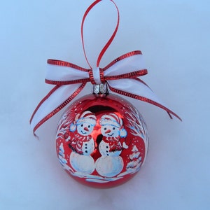 Puede incluir: Adorno navide&ntilde;o rojo con una cinta blanca y dos mu&ntilde;ecos de nieve pintados en la parte delantera. Los mu&ntilde;ecos de nieve llevan sombreros y bufandas.