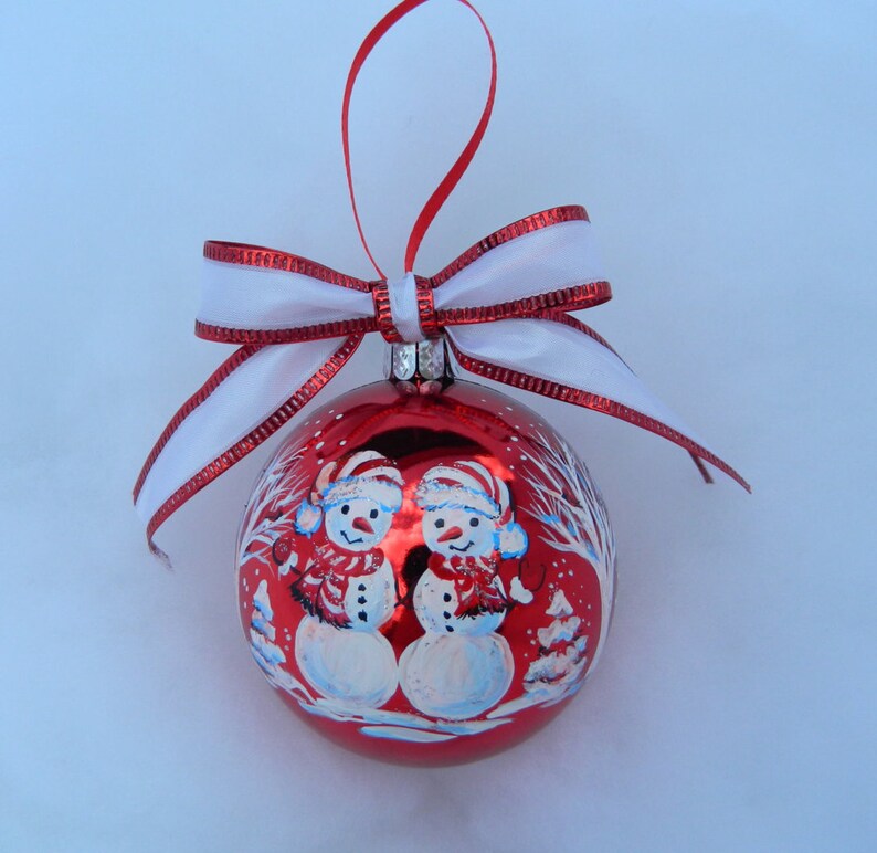 Puede incluir: Adorno navide&ntilde;o rojo con una cinta blanca y dos mu&ntilde;ecos de nieve pintados en la parte delantera. Los mu&ntilde;ecos de nieve llevan sombreros y bufandas.