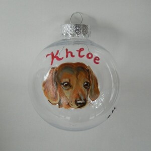 May include: Clear glass Christmas ornament with a hand-painted portrait of a brown and tan dachshund. The ornament is personalized with the name "Khloe" in red paint.