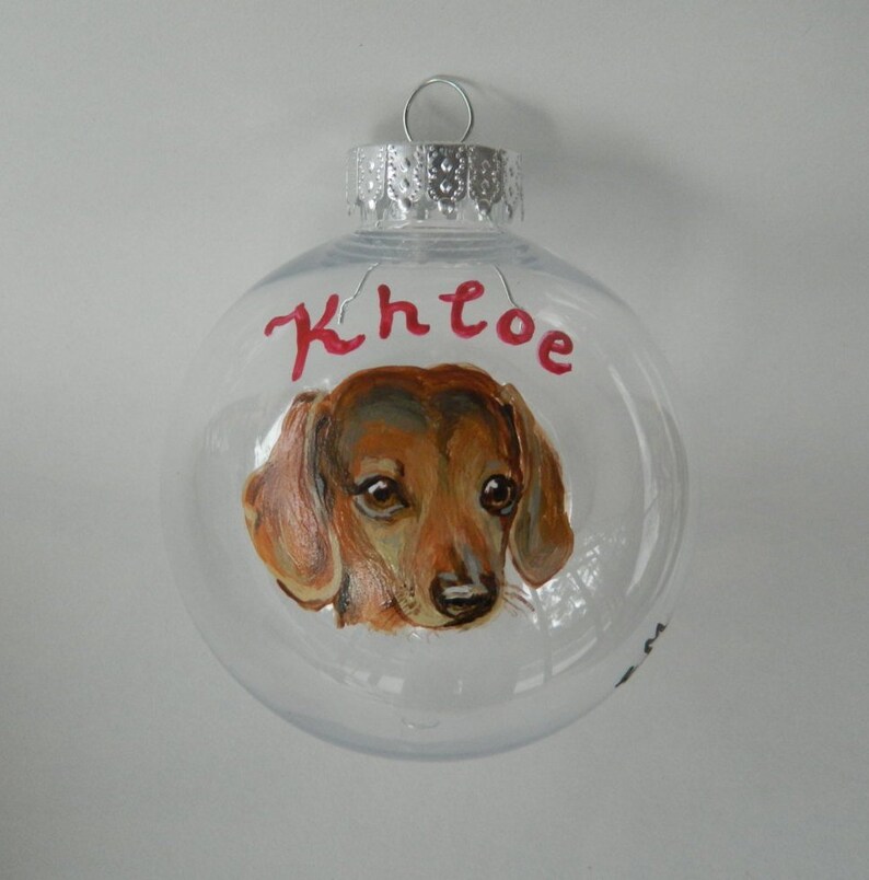May include: Clear glass Christmas ornament with a hand-painted portrait of a brown and tan dachshund. The ornament is personalized with the name "Khloe" in red paint.