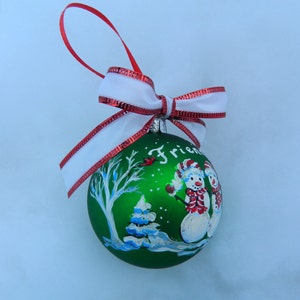 Puede incluir: Adorno navide&ntilde;o verde con una escena pintada de dos mu&ntilde;ecos de nieve, un &aacute;rbol y un cardenal. El adorno tiene una cinta blanca con ribete rojo y la palabra "Friends" est&aacute; pintada en la parte delantera.