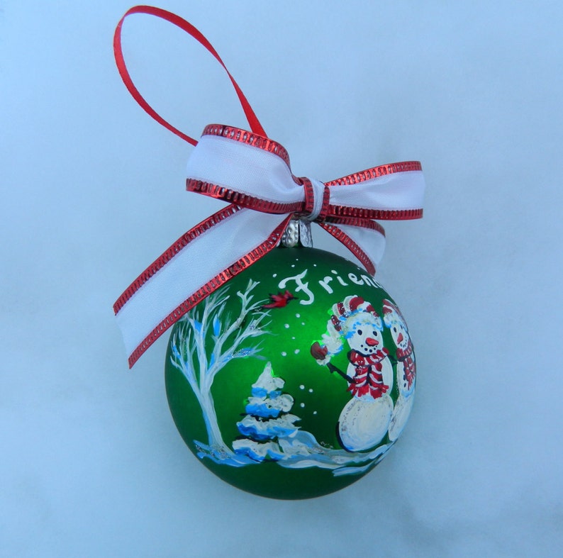 Puede incluir: Adorno navide&ntilde;o verde con una escena pintada de dos mu&ntilde;ecos de nieve, un &aacute;rbol y un cardenal. El adorno tiene una cinta blanca con ribete rojo y la palabra "Friends" est&aacute; pintada en la parte delantera.