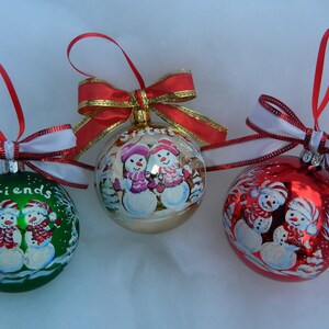 Puede incluir: Tres adornos navide&ntilde;os con dise&ntilde;os de mu&ntilde;ecos de nieve pintados a mano. Los adornos son rojos, verdes y dorados con cintas blancas. Los adornos tienen las palabras "Friends", "Sisters" y "Family" escritas en ellos.