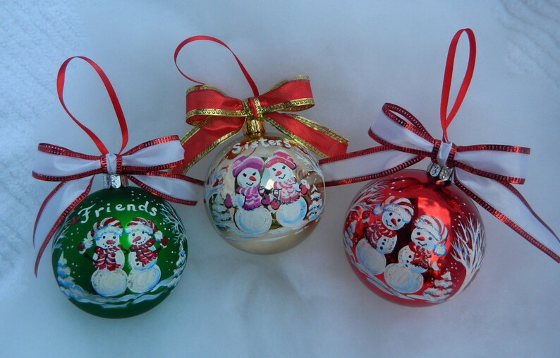 Puede incluir: Tres adornos navide&ntilde;os con dise&ntilde;os de mu&ntilde;ecos de nieve pintados a mano. Los adornos son rojos, verdes y dorados con cintas blancas. Los adornos tienen las palabras "Friends", "Sisters" y "Family" escritas en ellos.