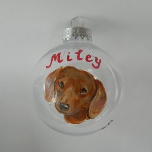 May include: Clear glass Christmas ornament with a hand-painted portrait of a brown dachshund dog. The ornament is personalized with the name "Miley" in red paint.