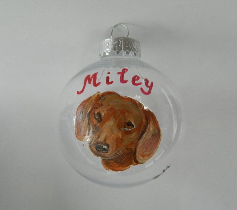 May include: Clear glass Christmas ornament with a hand-painted portrait of a brown dachshund dog. The ornament is personalized with the name "Miley" in red paint.