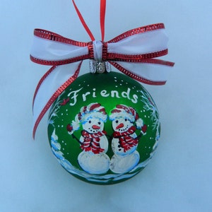 Puede incluir: Adorno navide&ntilde;o verde con un lazo blanco y ribete rojo. El adorno presenta una escena pintada de dos mu&ntilde;ecos de nieve con la palabra "Friends" escrita en la parte delantera.