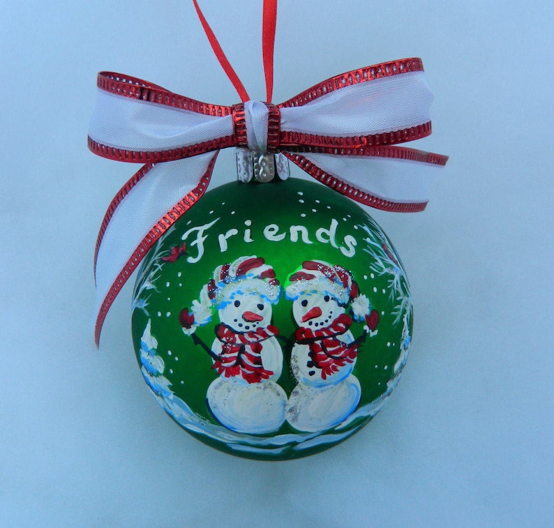 Puede incluir: Adorno navide&ntilde;o verde con un lazo blanco y ribete rojo. El adorno presenta una escena pintada de dos mu&ntilde;ecos de nieve con la palabra "Friends" escrita en la parte delantera.