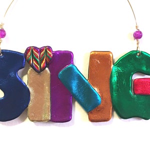 May include: A colorful clay ornament spelling out the word "SING" with a heart-shaped design in the middle of the "I". The ornament is strung on a gold wire with pink beads at the top.