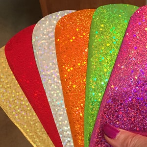 Könnte beinhalten: Ein Fächer aus schimmerndem Glitzerpapier in verschiedenen Farben, darunter Gold, Weiß, Rot, Orange, Grün und Pink. Jedes Blatt hat ein dezentes Sternenmuster.
