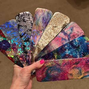 May include: A collection of eight iridescent vinyl sheets in various colors and patterns, including blue, pink, gold, and black. The sheets are arranged in a fan shape and have a holographic finish.
