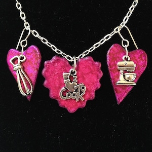 May include: A silver necklace with three pink heart charms. The center heart charm has a silver charm that says "I love to cook" with a small heart. The charms on the ends have a whisk and a stand mixer.
