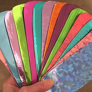 Op de afbeelding: Een waaier van kleurrijke, metallic en iriserende papierstalen. De stalen tonen een reeks kleuren, waaronder roze, blauw, groen en oranje. De papieren hebben een glanzend, reflecterend oppervlak en sommige hebben een textuur of patroon.