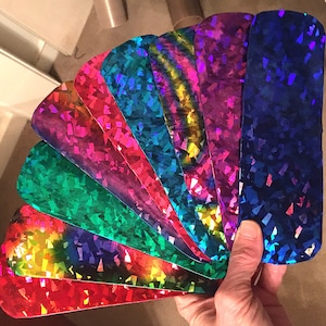 May include: A set of eight iridescent holographic sheets in various colors, including red, orange, yellow, green, blue, purple, and pink. The sheets are arranged in a fan shape and have a textured, glittery finish.