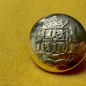 Op de afbeelding: Een zilverkleurige knoop met een gedetailleerd wapenontwerp, inclusief een kroon en diverse heraldische symbolen. De knoop is geplaatst tegen een gele, getextureerde achtergrond. Het ontwerp van de knoop is ingewikkeld en gedetailleerd.