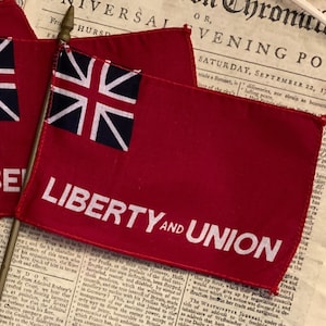 May include: Three miniature flags with red fabric and a white "LIBERTY" inscription. The flags feature a blue Union Jack design and are mounted on wooden sticks. The flags are displayed on a vintage newspaper with the text "UNIVERSAL EVENING POST" visible.