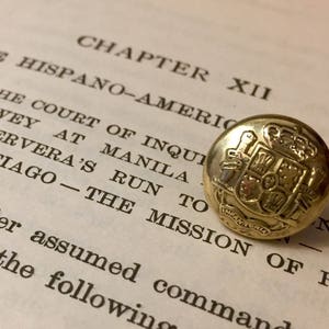 Könnte beinhalten: Ein goldfarbener Knopf mit einem detaillierten Wappen-Design. Der Knopf liegt auf einer Buchseite mit Text über den Spanisch-Amerikanischen Krieg.