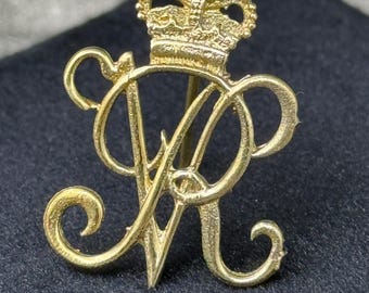 May include: A gold-colored brooch featuring the intertwined letters "R" and "A" beneath a detailed crown. The brooch has a pin on the back and is set against a dark background. The design is ornate and likely vintage.