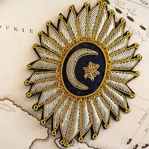 May include: A gold and silver embroidered patch with a crescent moon and star design on a dark blue background. The patch is surrounded by a circle of gold and silver rays.