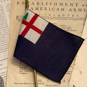 May include: A small, dark blue flag with a red cross and a green pine tree emblem, displayed on old paper documents. The flag's design suggests a historical or commemorative item, possibly a replica.