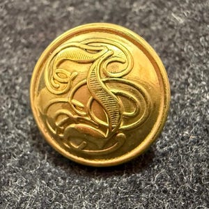 Pode incluir: Um botão dourado com um design em relevo. O botão tem formato circular com um padrão ornamentado em relevo no centro. Está sobre um fundo cinza escuro texturizado.