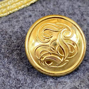 Pode incluir: Botão dourado com um design ornamentado apresentando uma letra "S" estilizada no centro. O botão é redondo e tem uma borda ligeiramente elevada. O botão está sobre um tecido cinza texturizado. O diâmetro do botão é de cerca de 2,5 cm.