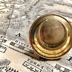 Puede incluir: Un sello de metal dorado con un diseño de media luna. El sello está apoyado en un mapa antiguo.