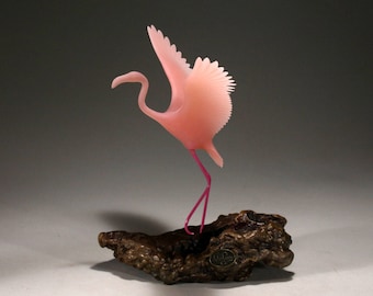 Pode incluir: Uma escultura de flamingo rosa de vidro com as asas abertas, em pé sobre um pedaço de madeira à deriva. A escultura é pintada à mão com detalhes rosa e roxo.