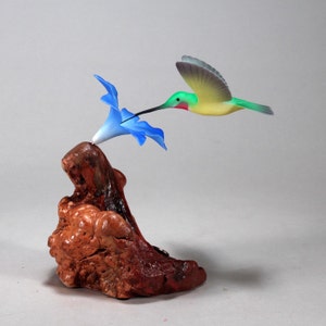 May include: A hand-carved wooden hummingbird sculpture perched on a piece of driftwood, with a blue flower. The hummingbird is painted green, yellow, and red, with black eyes. The sculpture is a realistic depiction of a hummingbird in flight.