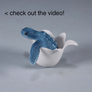 Może przedstawiać: Mała niebieska figurka żółwia wyłaniająca się z białego ceramicznego jajka. Żółw ma szczegółowy pancerz i realistyczny wygląd.