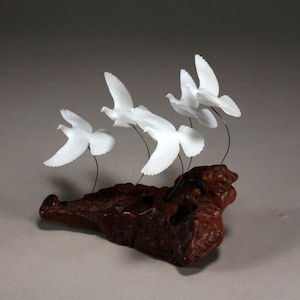 Op de afbeelding: Een sculptuur van vijf witte duiven in vlucht, geperst op een stuk drijfhout.