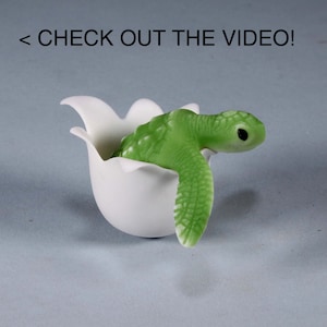 May include: A small green sea turtle figurine hatching from a white ceramic egg. The turtle has a detailed shell and a cute face.