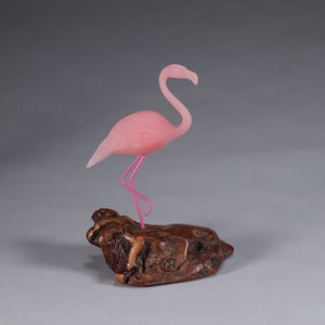 May include: A pink glass flamingo sculpture standing on a wooden base. The flamingo has pink legs and a long neck.