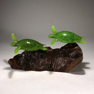 May include: Two green sea turtle sculptures on a wooden base. The turtles are in a swimming pose, with their heads and flippers extended. The base is made of dark brown wood with a natural grain.