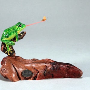 May include: A green frog figurine with a long pink tongue catching a yellow bee. The frog is perched on a brown wooden base.