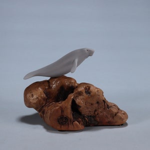May include: A white ceramic figurine of a manatee perched on a piece of driftwood. The figurine is detailed with realistic features and is a great addition to any marine life collection.