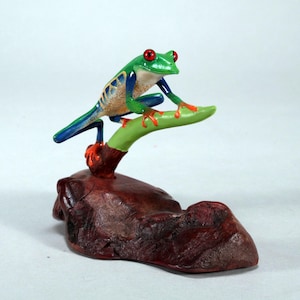 May include: A green and blue figurine of a red-eyed tree frog perched on a green branch, sitting on a brown wooden base.