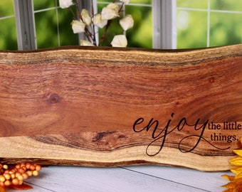 May include: A natural wood serving board with a live edge and a dark brown stain. The board has the words "enjoy the little things" engraved on it in a cursive font.