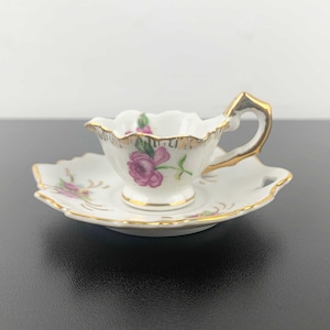 May include: A miniature white porcelain teacup and saucer set with a floral design. The cup has a gold rim and a delicate handle. The saucer has a scalloped edge.