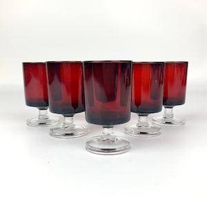 Puede incluir: Juego de seis vasos de chupito de vidrio rojo con tallos y bases de vidrio transparente. Los vasos están dispuestos en semicírculo sobre una superficie blanca.
