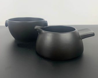 Op de afbeelding: Een zwarte keramische roomkan en suikerpot set. De roomkan heeft een rond lichaam en een plat handvat. De suikerpot heeft een rond lichaam en twee kleine handvatten.