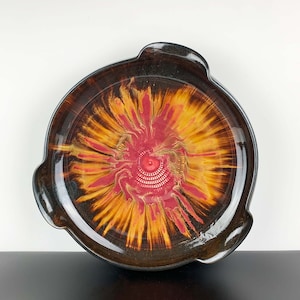 Op de afbeelding: Een ronde keramische serveerschaal met een donkerbruine glazuur en een rood en geel swirl-ontwerp in het midden.