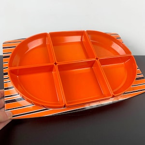 May include: An orange serving tray with six compartments. The tray is set on a rectangular base with orange and black stripes. The tray is made of a glossy material and is designed for serving snacks or appetizers.
