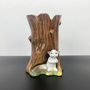 Puede incluir: Un jarrón de cerámica con forma de tronco de árbol con una figurita de ardilla blanca sentada en la base. El jarrón es marrón con una superficie texturizada y la ardilla tiene una cola marrón claro y ojos negros.