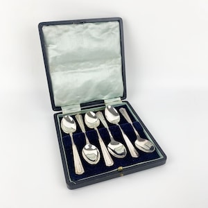 May include: A set of six silver-toned spoons with tapered handles arranged in a dark blue velvet-lined presentation box. The box features a hinged lid with a pale blue satin interior lining and a secure metal clasp closure on the front edge.