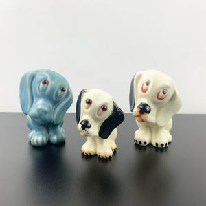 May include: Three ceramic dog figurines. One is blue, one is white with black ears, and one is white with black ears and a black nose.