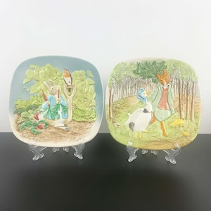 Puede incluir: Dos platos de pared de cerámica con personajes de los libros infantiles de Beatrix Potter. El primer plato representa a Peter Rabbit comiendo una zanahoria, mientras que el segundo plato muestra al Sr. Fox y al ganso.