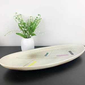 May include: An oval ceramic serving platter in off-white with a speckled finish. It has abstract, colourful accents including yellow, blue, purple, and grey shapes. A small white vase with white flowers is in the background.